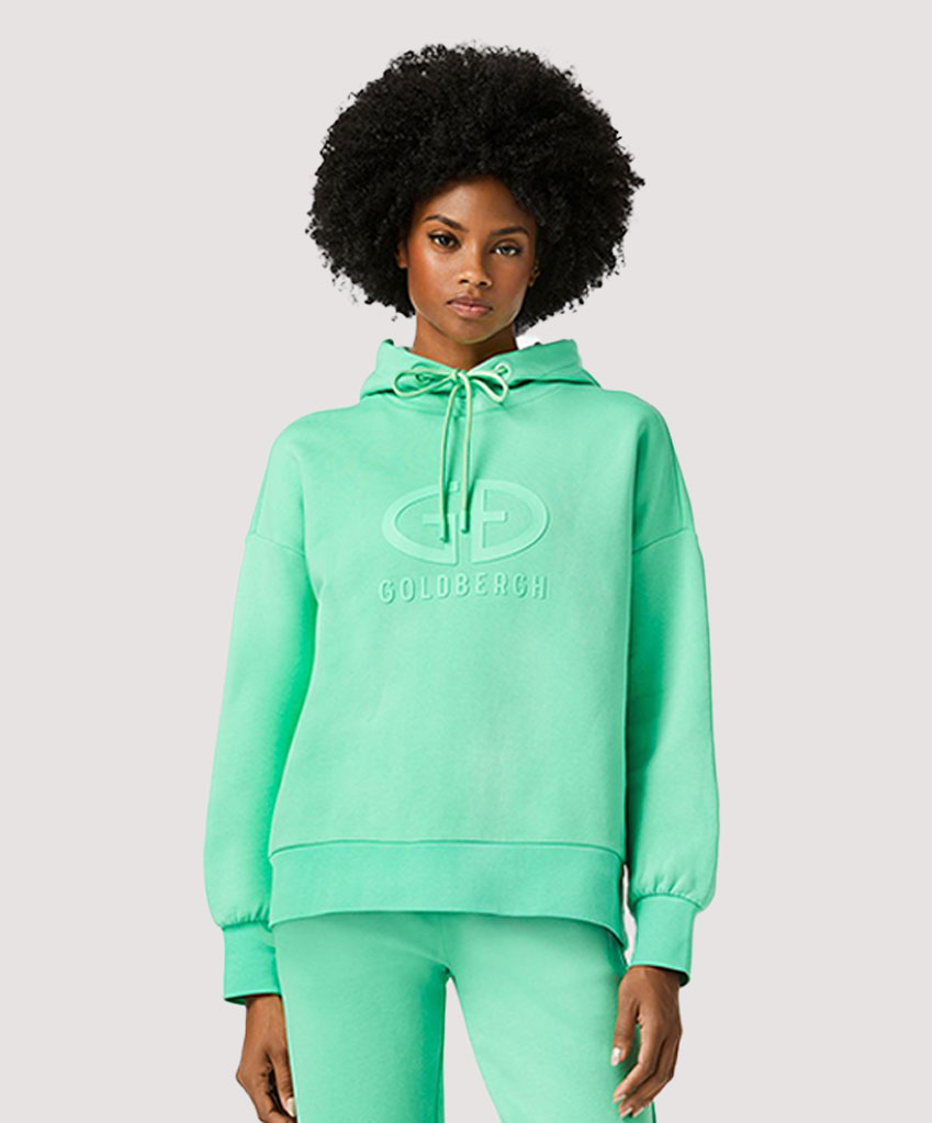 Een getinte dame in een groene goldbergh sweater, sweatpants en witte schoenen. Model voor op de homepage van Ab Geldermans Sport